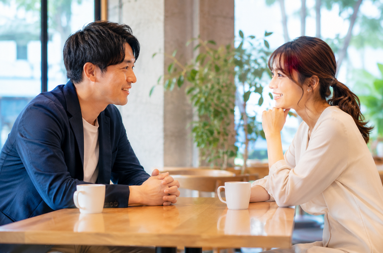 カフェのテーブル越しに向かい合う20代女性と30代男性。二人とも笑顔でリラックスした雰囲気。コーヒーカップが置かれた明るい空間。