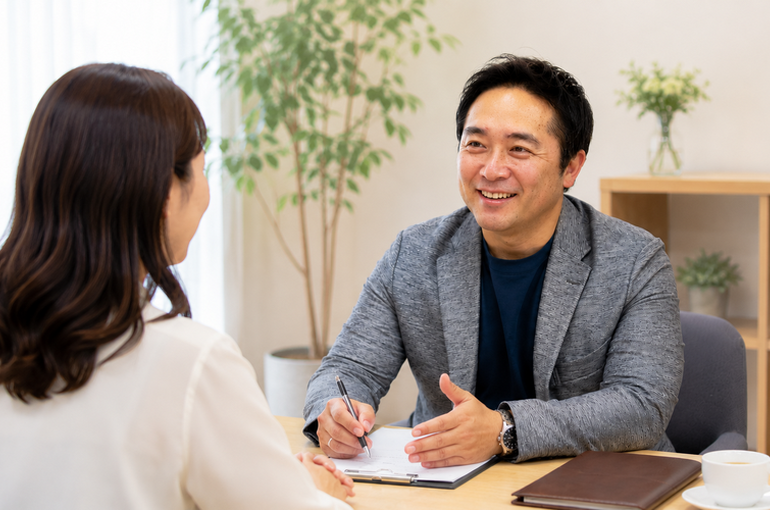 婚活カウンセリングで笑顔で相談に乗る男性コンサルタントと、真摯に耳を傾ける女性。屋内のオフィスで行われる相談風景。