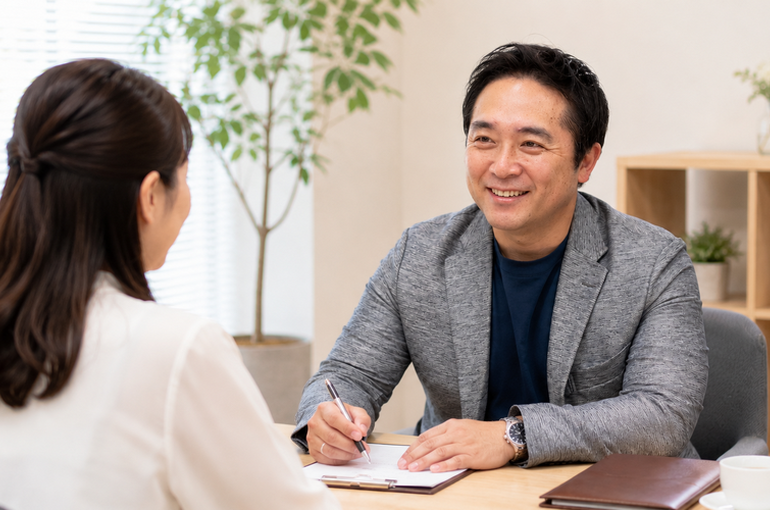 グレーのジャケットを着た40代男性がメモを取りながら笑顔で女性と面談している、明るいオフィス空間での婚活カウンセリングの様子
