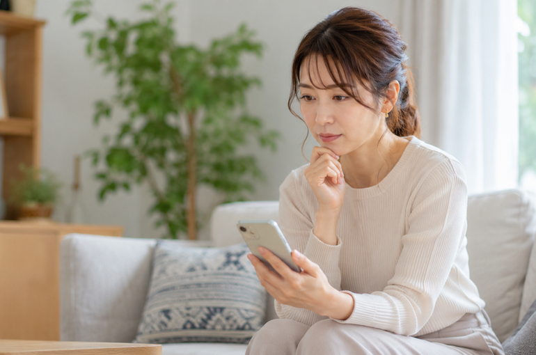 スマートフォンを持ちながら考え込む30代女性が、明るいリビングのソファに座っている様子を捉えた写真で、婚活での悩みや感情表現について思考している場面